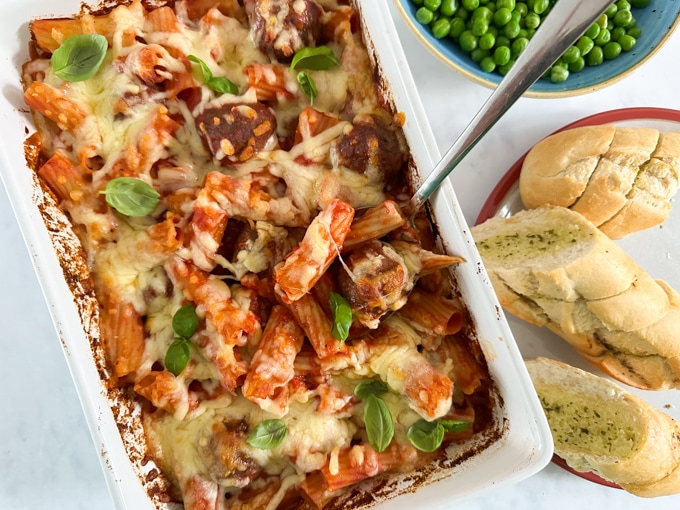 Easy meatball pasta bake fresh out of the oven in a large rectangle oven dish, accompanied with green peas and crusty garlic bread slices. 