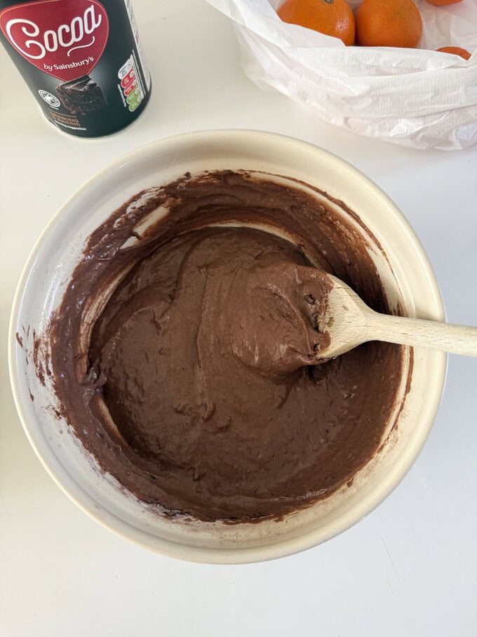 Ingredients  being added to a cream colored mixing bowl. and being stirred with a wooden spoon.